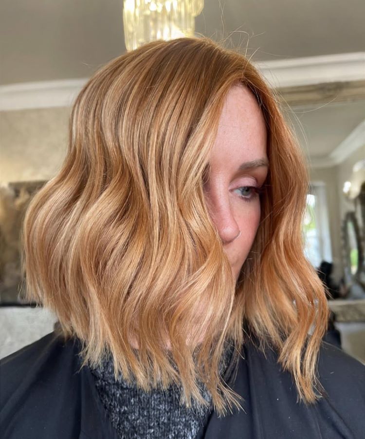 Mulher com cabelo médio e ondulado, destacando uma das cores de cabelo tendência: loiro acobreado com nuances quentes e brilhantes. Ela veste roupa preta com gola cinza e está em um ambiente interno com detalhes elegantes e luz suave.





