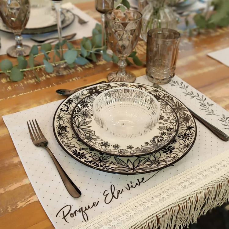 Uma mesa de jantar decorada para a Páscoa Cristã: nela contém pratos, talheres, copos, taças e enfeites. O sousplat é branco com detalhes em renda e tiras com a frase "Porque Ele Vive". As cores predominantes são branco, marrom, verde e preto.