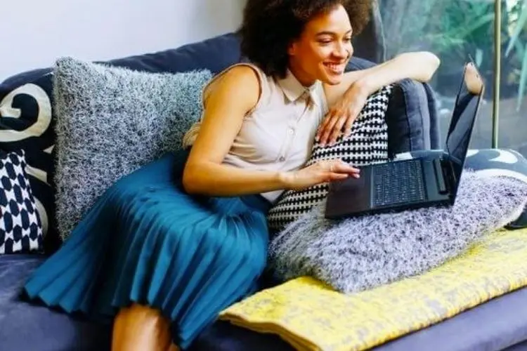 Mulher de pele negra usando regata bege e saia midi azul plissada, deitada em sofá azul com almofadas e mexendo em notebook