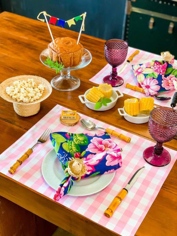A imagem representa decoração para festa junina. Contém bolo, milho, pipoca, paçoca, em mesa para dois, com jogo americano xadrez