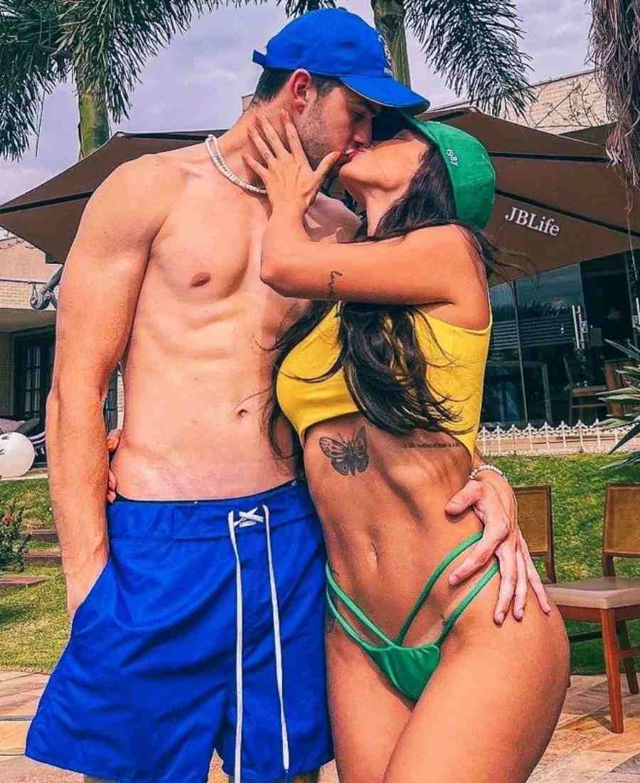 Imagem com fundo de área de piscina. No centro, casal usando roupa de banho copa do mundo: short azul masculino + boné azul. Top amarelo + biquíni verde + boné verde.