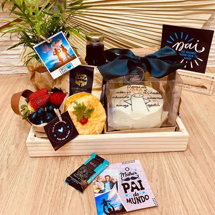 A imagem mostra uma cesta de Dia dos Pais com diversos itens. Há uma garrafa, frutas, chocolates e um cartão com a frase “O melhor pai do mundo”. A cesta é decorada com um laço azul e tem ao fundo uma planta e um cartão-postal.

