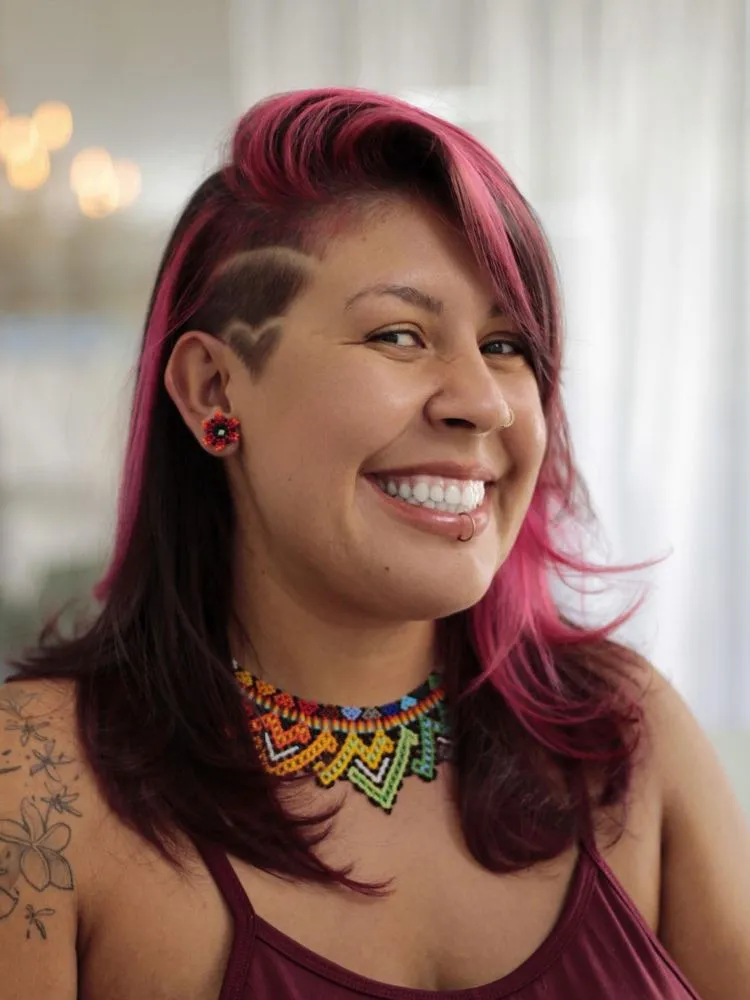 Mulher de pele morena e rosto redondo sorrindo, usando sidecut com coração no cabelo médio rosa