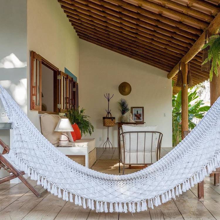 Imagem de fachada de casa simples, com alpendre, que mostra uma casa branca com sofá, poltronas, rede branca