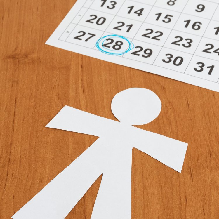 A imagem mostra um calendário com uma folha destacando os dias do mês. O dia 28 está circulado em azul. Em primeiro plano, há uma figura recortada de papel em forma de pessoa sobre uma superfície de madeira. A cena sugere uma conexão com uma data importante, possivelmente relacionada a pessoas ou eventos significativos.


