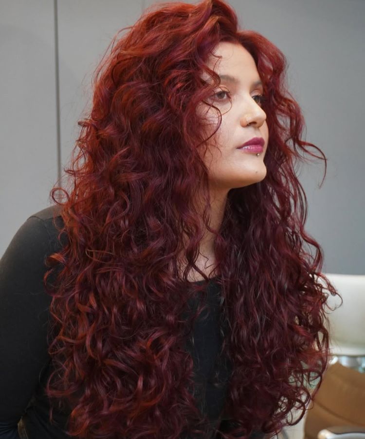 Mulher com cabelos longos, volumosos e cacheados, exibindo cores de cabelo em tom vermelho intenso e iluminado. Ela veste uma blusa preta, usa batom rosado, possui um piercing no nariz e está em um ambiente interno com fundo neutro.






