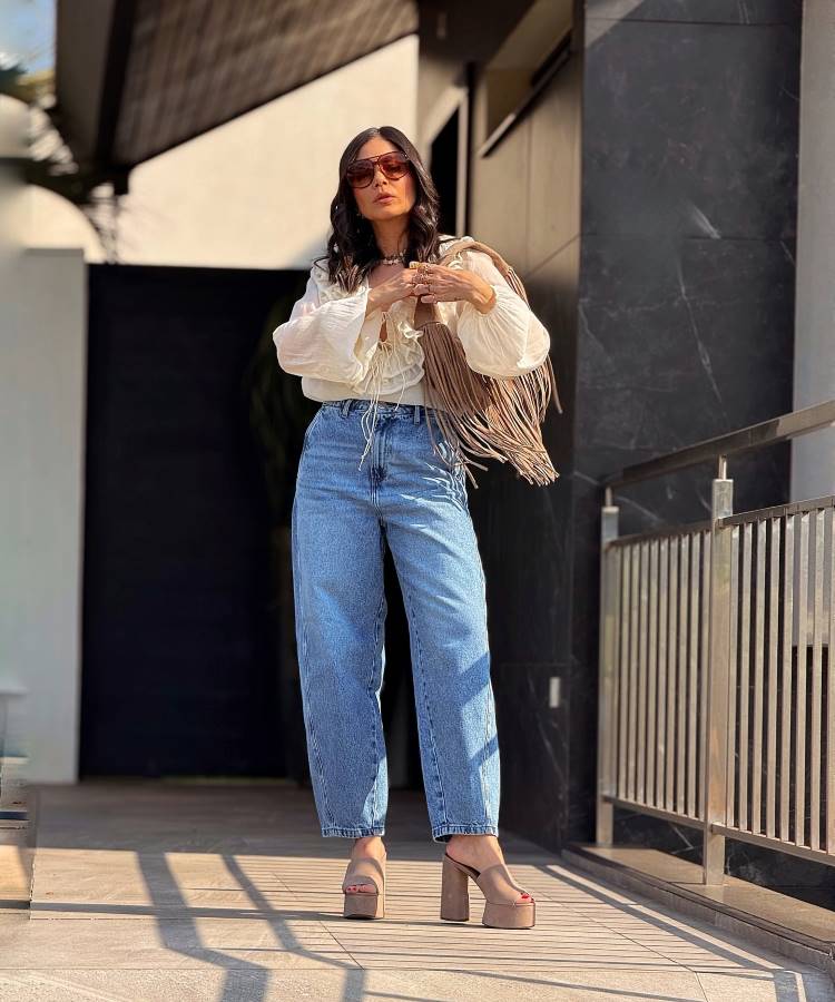 A imagem apresenta uma mulher usando uma calça jeans reta de lavagem clara, tendência 2025, com cintura alta e caimento confortável. Ela combina a peça com uma blusa fluida branca, bolsa de franjas e sandálias plataforma, criando um visual boho-chic moderno.