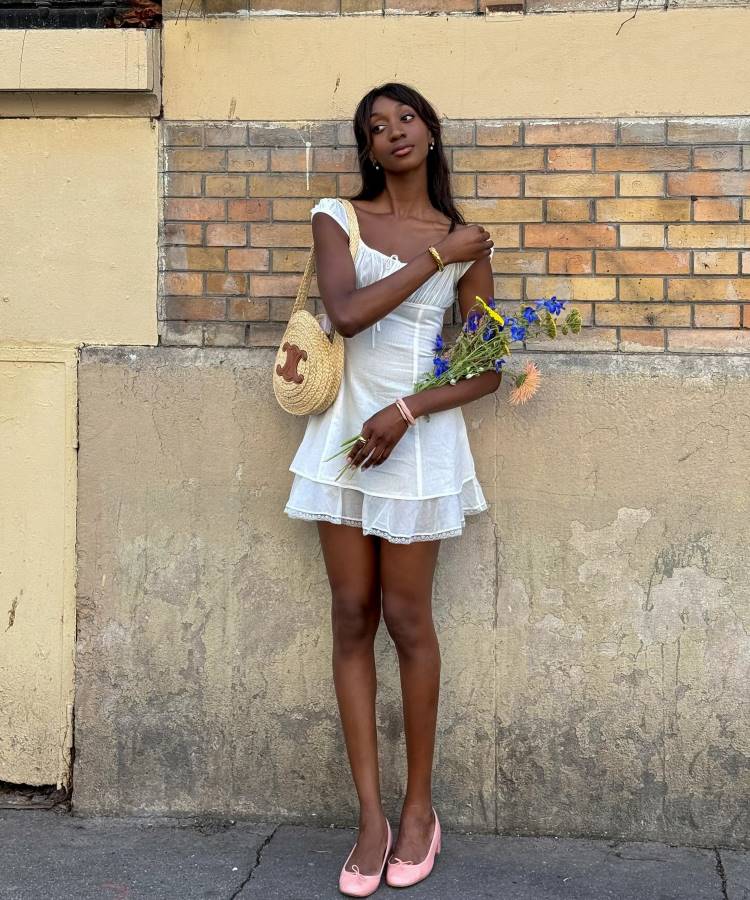 Uma mulher magra e alta, com longos cabelos escuros e um vestido de alças branco, posa em frente a uma parede de tijolos. Ela segura um buquê de flores azuis e exibe tendências do verão europeu, como sua bolsa de palha e sapatilhas rosas.