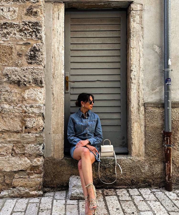 Uma mulher com cabelo escuro e óculos de sol senta-se em uma entrada de pedra. Ela veste um vestido jeans, amarra as sandálias e tem uma bolsa branca ao lado dela, olhando para o lado.







