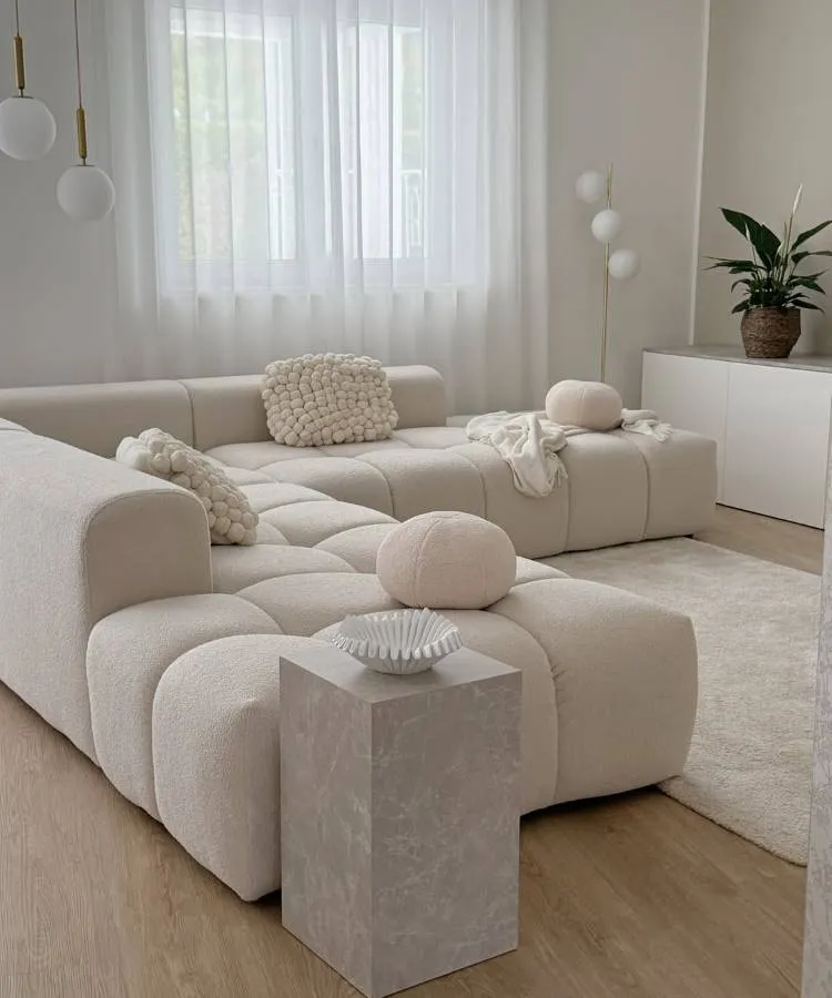 Uma sala de estar minimalista com sofá modular bege claro e almofadas para sala texturizadas e redondas. Uma mesa lateral quadrada de mármore claro exibe um bowl branco. Cortinas brancas translúcidas e uma luminária de piso completam o ambiente suave e sereno, com um toque de verde de uma planta.







