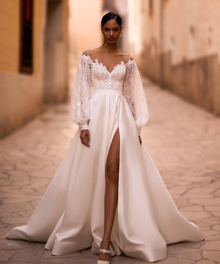 Noiva em vestido branco elegante com corpete rendado, mangas bufantes transparentes e saia fluida com fenda alta. O design sofisticado combina delicadeza e modernidade, enquanto o cenário de rua de pedra adiciona um charme rústico e atemporal.





