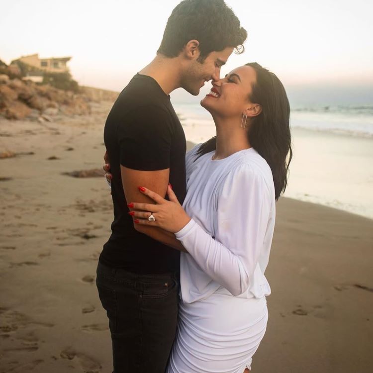 Demi Lovato e Max Ehrich sorridente em pé na praia ao entardecer, tocando narizes. Ela usa um vestido branco com anel destacado na mão, e ele veste camiseta preta. Ao fundo, areia, água e um tom suave de céu.





