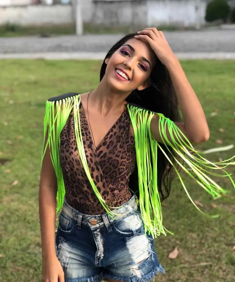 A mulher sorridente usa um body estampado de oncinha, shorts jeans rasgados e ombreiras neon com franjas verdes vibrantes. Seu cabelo longo está solto, e ela toca a cabeça com uma mão. O fundo possui grama e calçada.