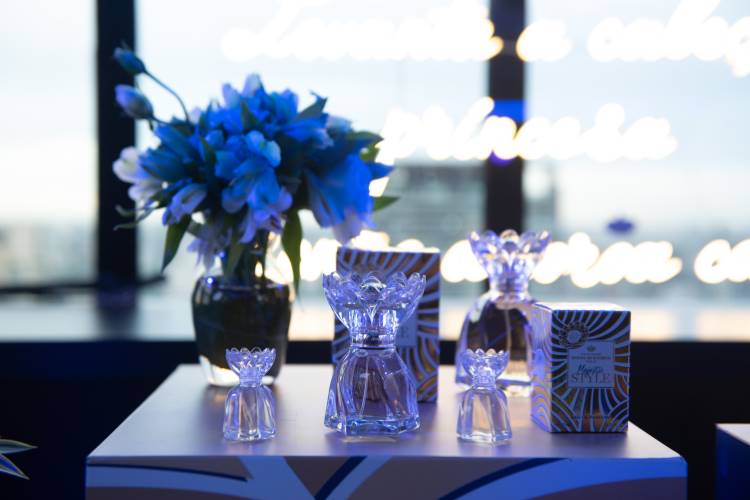 Frascos de perfume cristalinos com tampas decorativas em uma mesa azul e dourada. Embalagens ao fundo e um vaso de flores azuis completam a cena. A iluminação suave destaca os produtos, criando uma atmosfera elegante e sofisticada no ambiente.