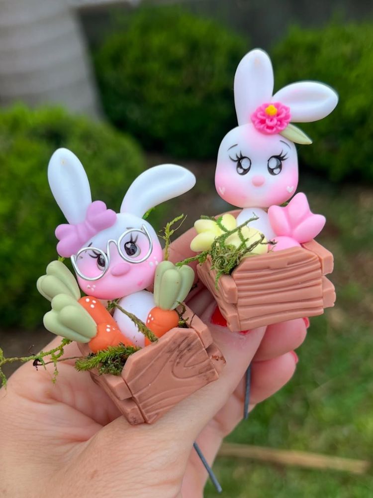 Mão de pele clara segurando coelhos de biscuit em caixas, uma de cenoura e outra de ovos coloridos