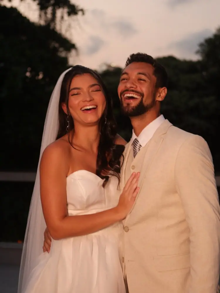 Lara Santana com vestido de noiva sem alça, véu, abraçada com Matheus, que usa terno bege 