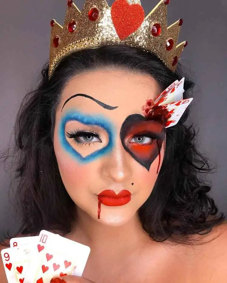 Mulher de pele clara usando maquiagem de Rainha de Copas, com coração azul em um olho e coração vermelho e preto com cartas enfiadas na pele em outro olho