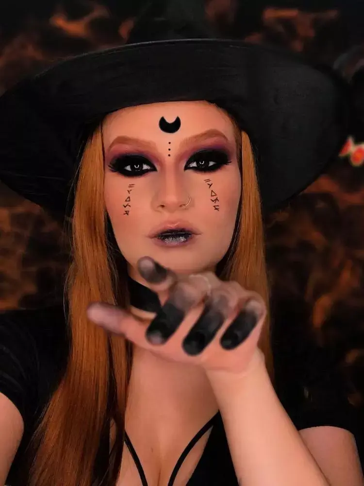 Mulher ruiva de pele clara fantasiada de bruxa com dedos pretos, lua preta na testa, desenhos misticos na bochecha