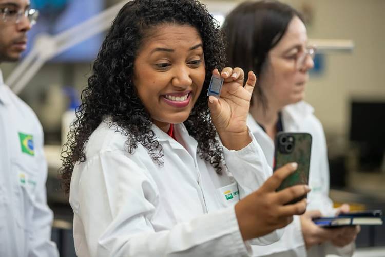 Mulher sorridente, vestindo jaleco branco, segura um chip enquanto grava conteúdo com celular. Ao fundo, outros profissionais em laboratório. Representa influencers de educação que compartilham ciência e tecnologia nas redes sociais de forma acessível e inspiradora.