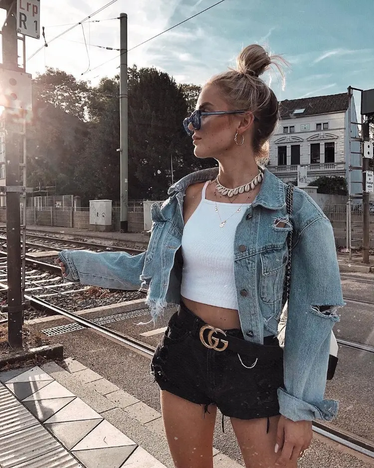 Frase para foto pensativa mulher loira usa coque, óculos de sol gatinho, choker de búzios, cinto preto Gucci e veste short jeans destroyed. camisa branca e jaqueta jeans curta Frase para foto pensativa