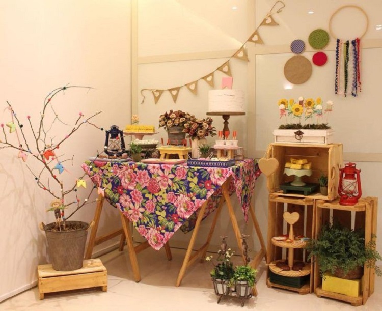 A imagem representa decoração para festa junina com mesas diferentes. Contém doces, bandeirinhas, arranjos de flores, lampião, etc
