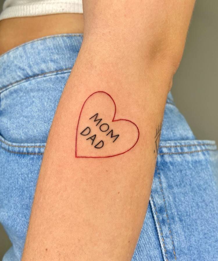 A imagem mostra uma tatuagem no braço de uma pessoa. É um contorno de coração em linha fina vermelha, com as palavras "MOM" e "DAD" escritas em preto dentro dele. A pessoa veste calça jeans azul, e a pele é clara.







