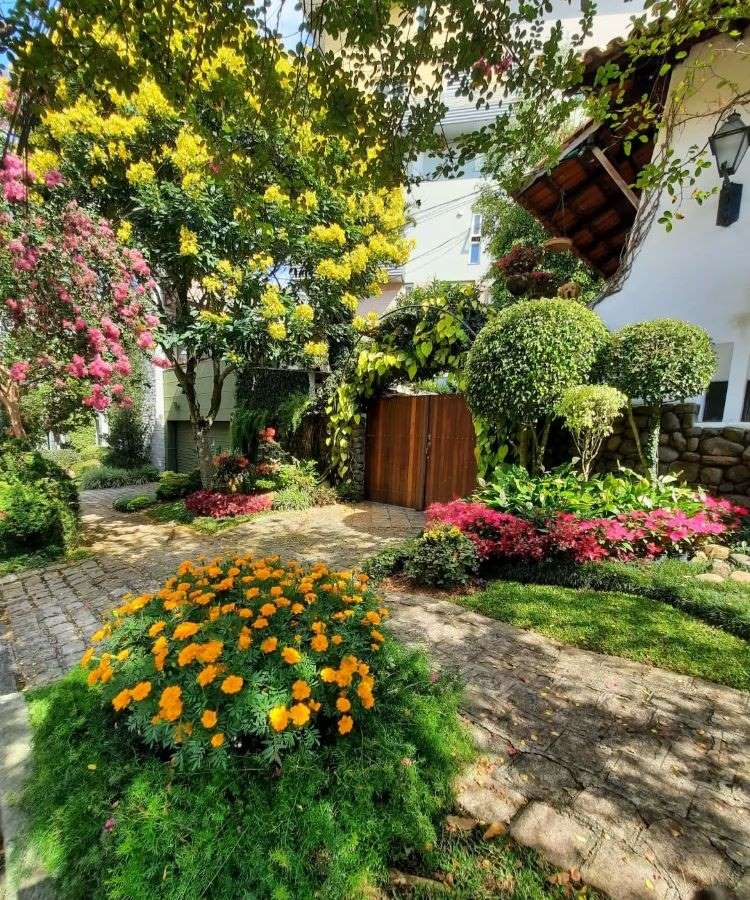 A imagem mostra um jardim vibrante com flores amarelas, rosas e alaranjadas em meio a folhagens verdes. Árvores sombreadas cercam um caminho de pedras que leva a um portão de madeira, compondo um cenário acolhedor e harmonioso.