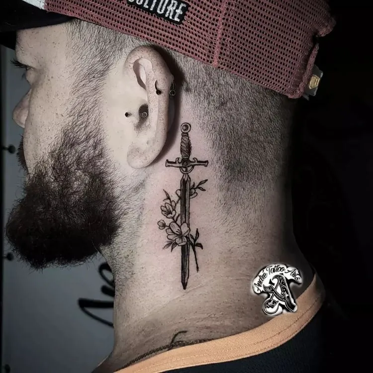 A imagem mostra uma pessoa de perfil usando um boné vermelho e preto. Ela tem uma tatuagem no pescoço de uma espada vertical cercada por flores. A pessoa tem barba e usa vários piercings na orelha.


