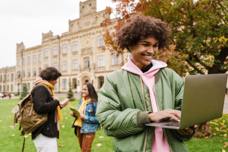 Jovem sorridente usando laptop em um campus universitário ao ar livre, com grama e árvores ao fundo. Duas pessoas conversam próximas, uma segurando um celular. Edifício histórico com arquitetura clássica completa o cenário iluminado por luz natural. Ele utiliza uma ferramenta de paráfrase para garantir a autenticidade do conteúdo.