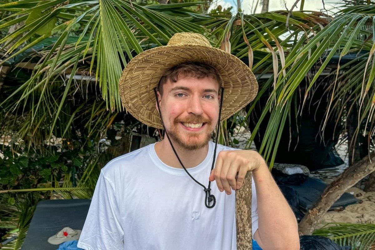 A imagem mostra MrBeast sorridente usando um chapéu de palha e camiseta branca, em meio a uma vegetação tropical com folhas de palmeiras ao fundo. Ele segura um objeto de madeira, transmitindo um clima descontraído. Ilustra quanto ganham os 10 maiores influenciadores do mundo.