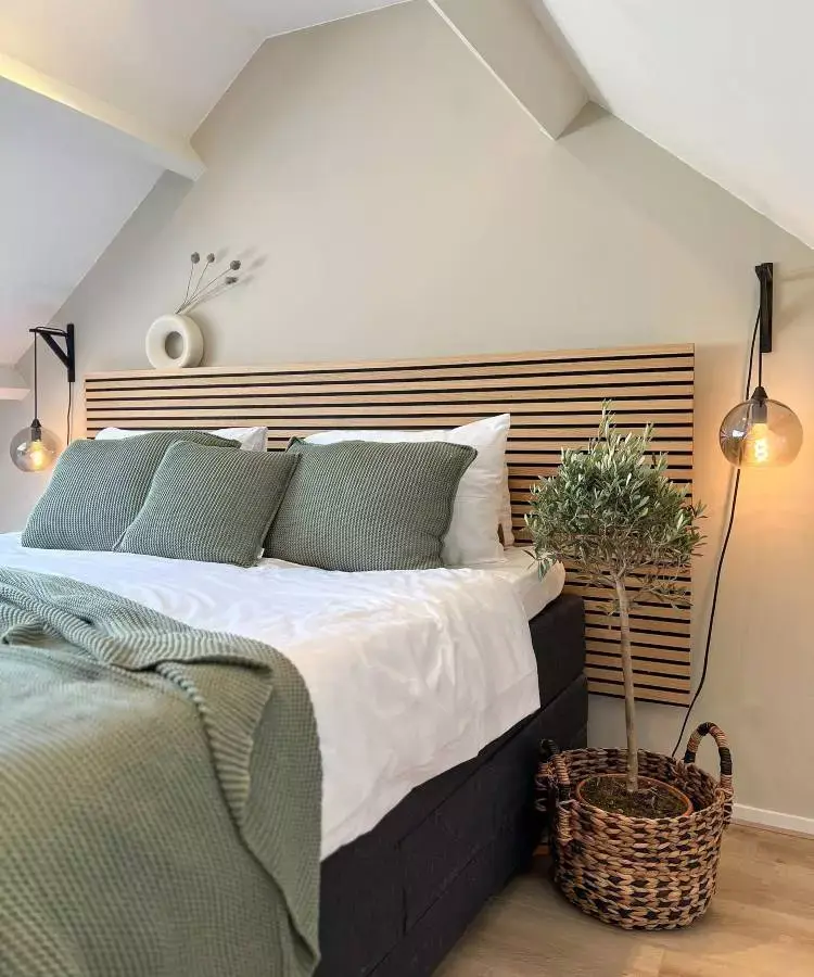 Quarto de casal com roupa de cama verde e branco, cabeceira de madeira e planta no canto da cama + luminárias penduradas