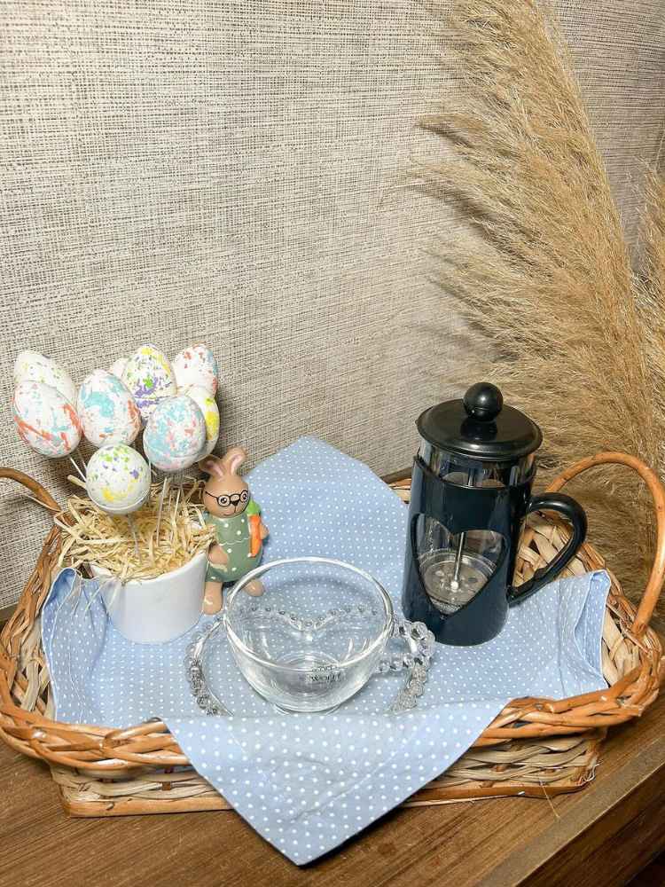 Cantinho do café com prensa francesa preta, arranjo de ovos de Páscoa coloridos, xícara e pires transparentes, tudo dentro de cesta de palha com toalha azul.