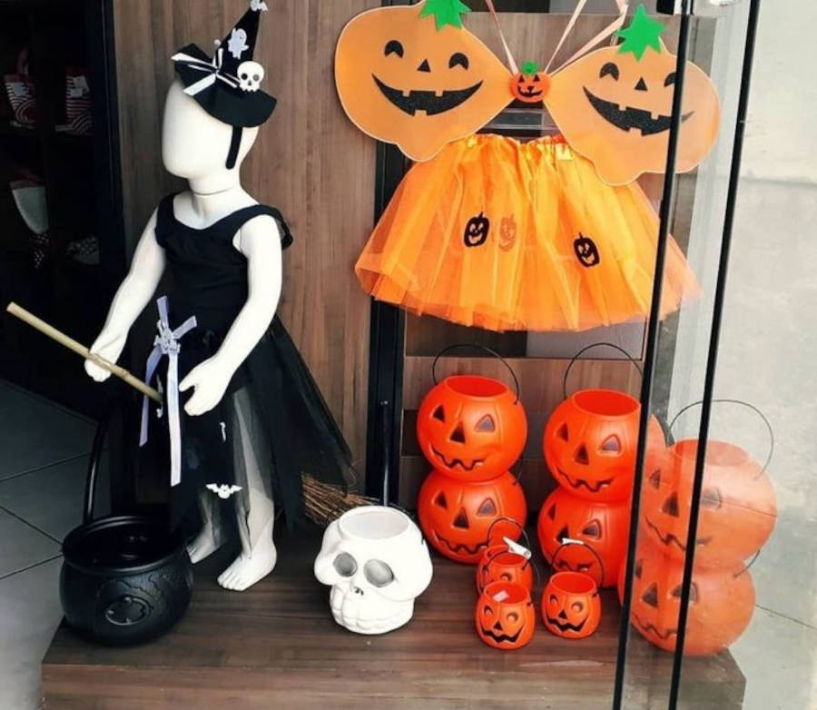 Foto de vitrine  infantil enfeitada para o Halloween, com manequimd e criança vestido de bruxa e enfeites de abóbora e caveira