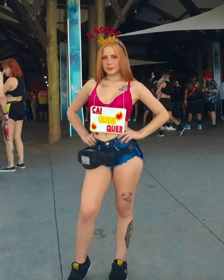 Mulher de cropped rosa, shorts jeans, tiara escrito "golpe tá aí" e placa escrito "cai quem quer"