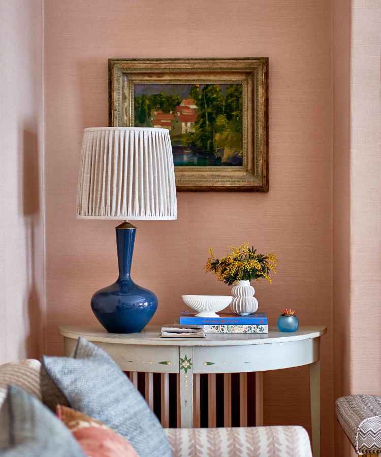 A imagem mostra um canto aconchegante com parede rosa, mesa vintage azul-clara decorada com abajur azul, livro, vaso de flores amarelas e pequena escultura. Um quadro com paisagem e sofá com almofadas complementam a atmosfera acolhedora e elegante. - tendências de decoração para 2025