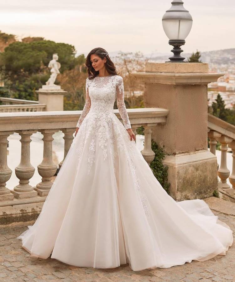 Noiva em vestido de noiva clássico com mangas longas rendadas, corpete bordado e saia volumosa de tule, destacando as tendências em vestidos de noiva 2025. O cenário ao ar livre, com balaústres e estátuas, acrescenta um toque romântico e sofisticado.