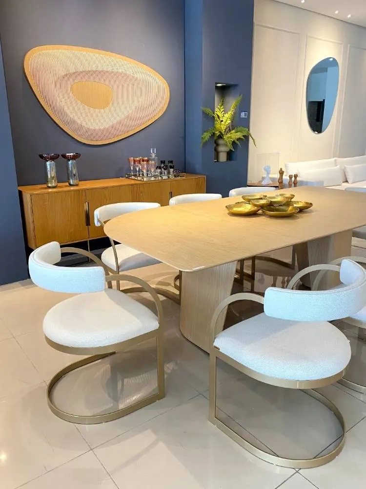 Sala de jantar com parede destacada azul, móvel em madeira, espelho orgânico, mesa e cadeiras arredondadas. 