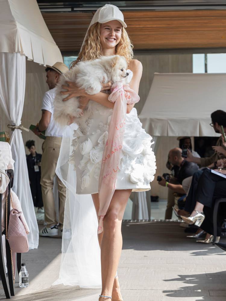 Mulher loira de pele clara usando vestido de noiva curto com flores, boné e segurando Lulu da Pomerânia no colo