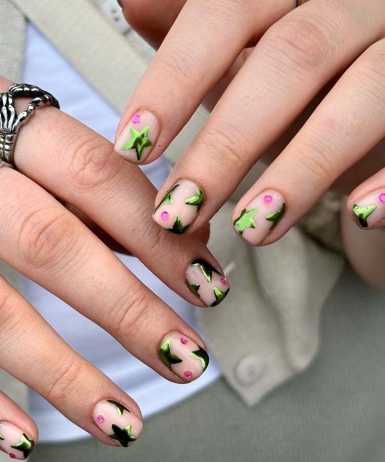 Mãos com unhas decoradas em base nude apresentando estrelas verdes brilhantes e pequenos pontos rosa vibrantes. A luz natural destaca os detalhes. Um anel prateado com design de esqueleto adorna o dedo. Fundo com roupas em tons neutros desfocado.