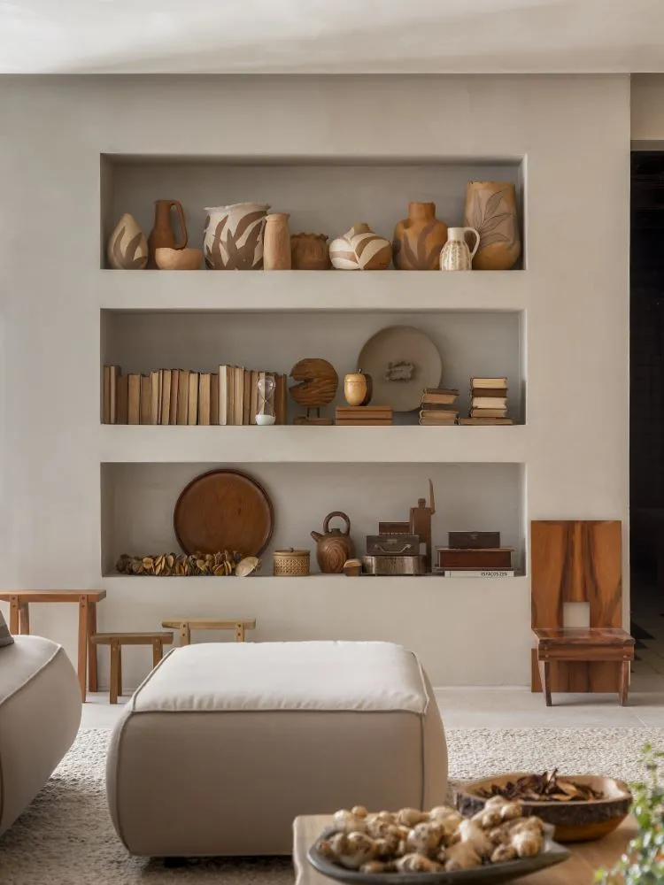 Decoração de estante de uma sala de estar em tons neutros, com puff e diversos vasos de cerâmica e itens decorativos em tons terrosos, que contribui com a saúde mental.