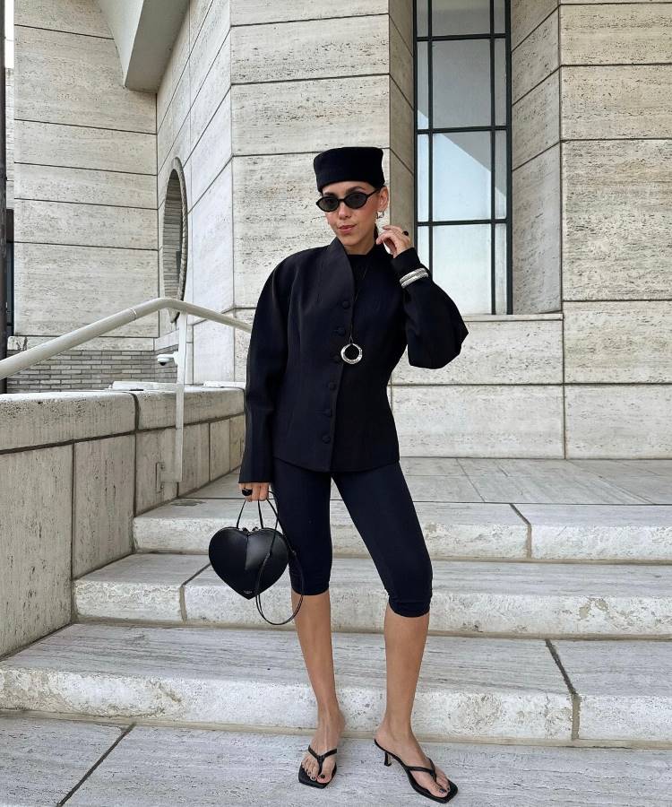 Uma mulher com chapéu preto, óculos de sol e bolsa em forma de coração posa em uma escadaria de pedra. Ela veste um conjunto preto de calça capri, blazer elegante com mangas bufantes e chinelos de salto.