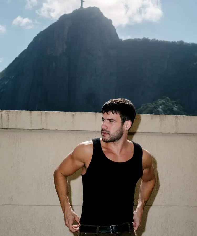 Homem musculoso de regata preta posa com o Corcovado, montanha no Rio de Janeiro, ao fundo. A estátua do Cristo Redentor é visível no topo, sob a luz do sol.