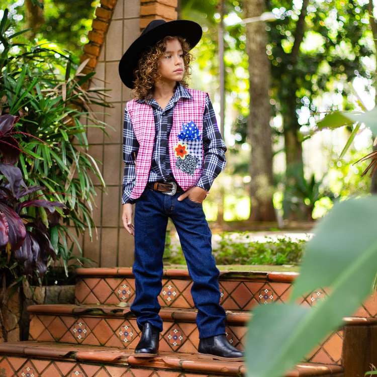 Menino usando camisa xadrez com colete, calça jeans, chapéu e bota, look country.

