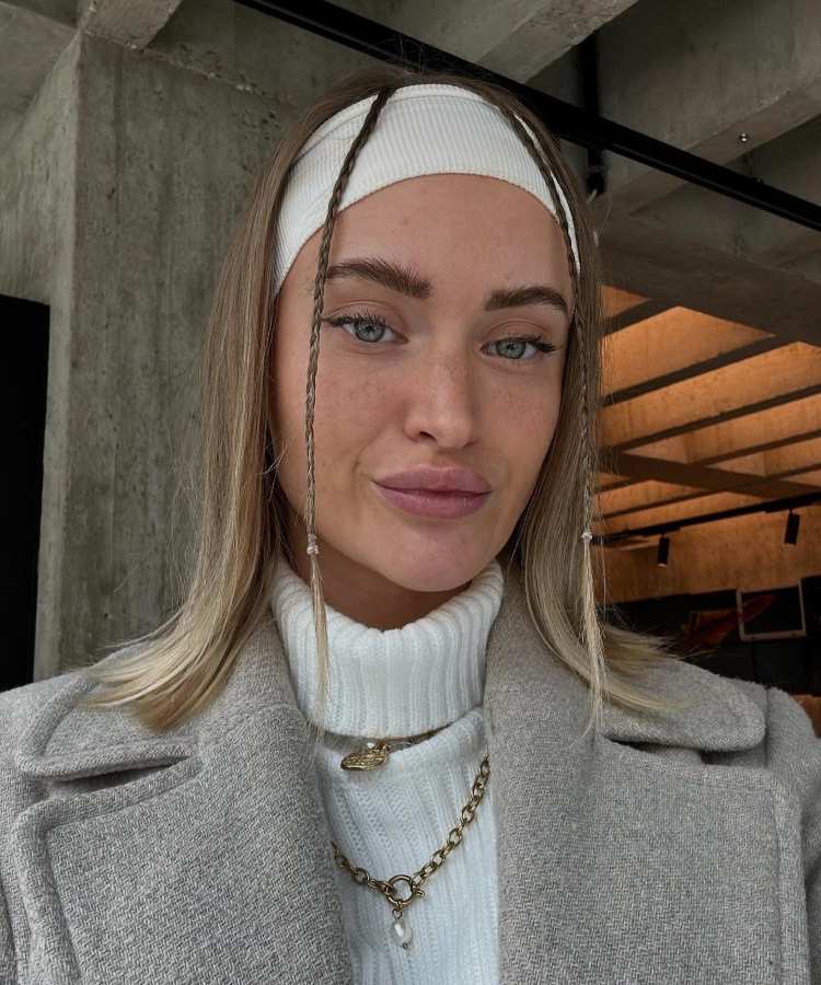 Mulher com cabelo loiro liso, dividido ao meio, fios frontais trançados, faixa branca, usando casaco cinza e suéter branco, posando em ambiente moderno.
