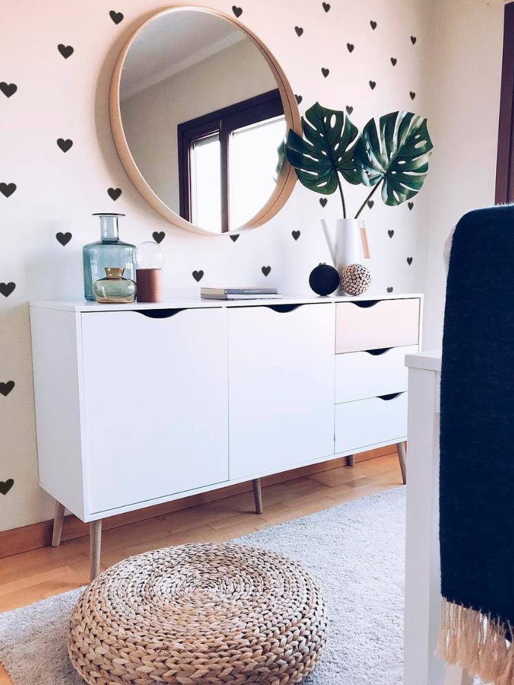 Aparador branco em frente a tapete bege, com vaso de planta e outros itens decorativos, além de espelho e, atrás, parede branca com adesivos de corações pretos