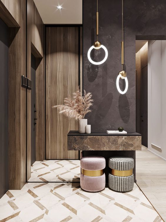 Hall de entrada com paredes escuras e de madeira, aparador suspenso com livro, vela e vaso com planta, luminárias circulares pendentes e puffes, um bege e outro branco e preto, ambos com detalhe em doruado 