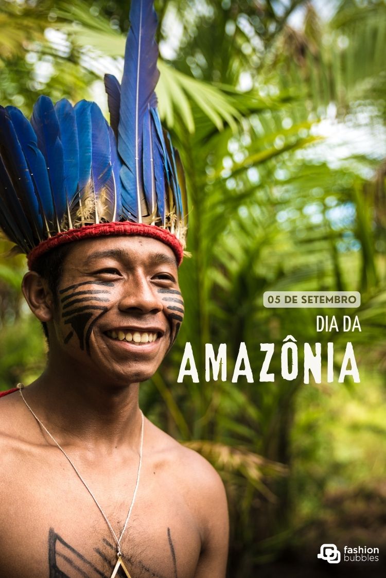 Um homem indígena sorridente com pintura facial e um cocar de penas azuis vibrantes posa contra uma floresta verde embaçada. O texto à direita diz "05 DE SETEMBRO DIA DA AMAZÔNIA", promovendo o Dia da Amazônia. A imagem evoca orgulho cultural e beleza natural.