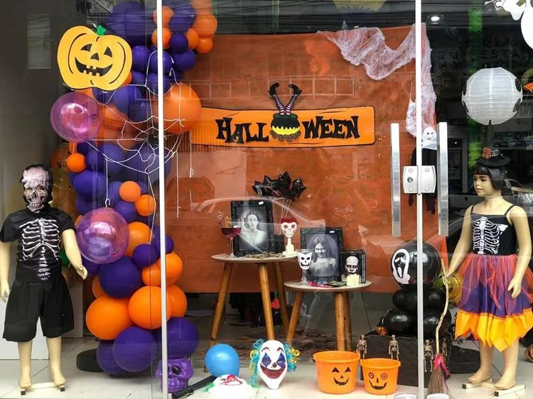Vitrine de Halloween com painel laranja, teias, bexigas em laranja e roxo, dois manequins com fantasias de esqueleto, mesas com retratos em branco e preto, baldes de abóbora e mais 