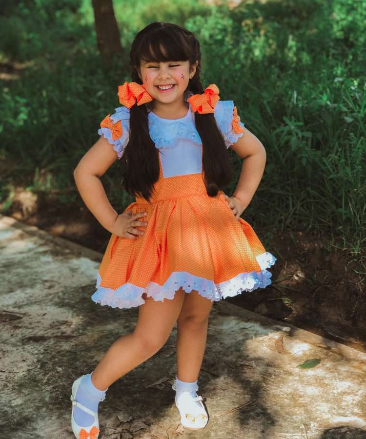 Menina com vestido de festa junina branco e laranja, de bolinha. No cabelo, laços grandes laranja