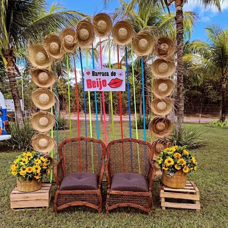 Um cenário de festa Junina ao ar livre com um arco de chapéus de palha e fitas coloridas. Duas cadeiras de vime com almofadas escuras e cestas de girassóis completam a decoração.